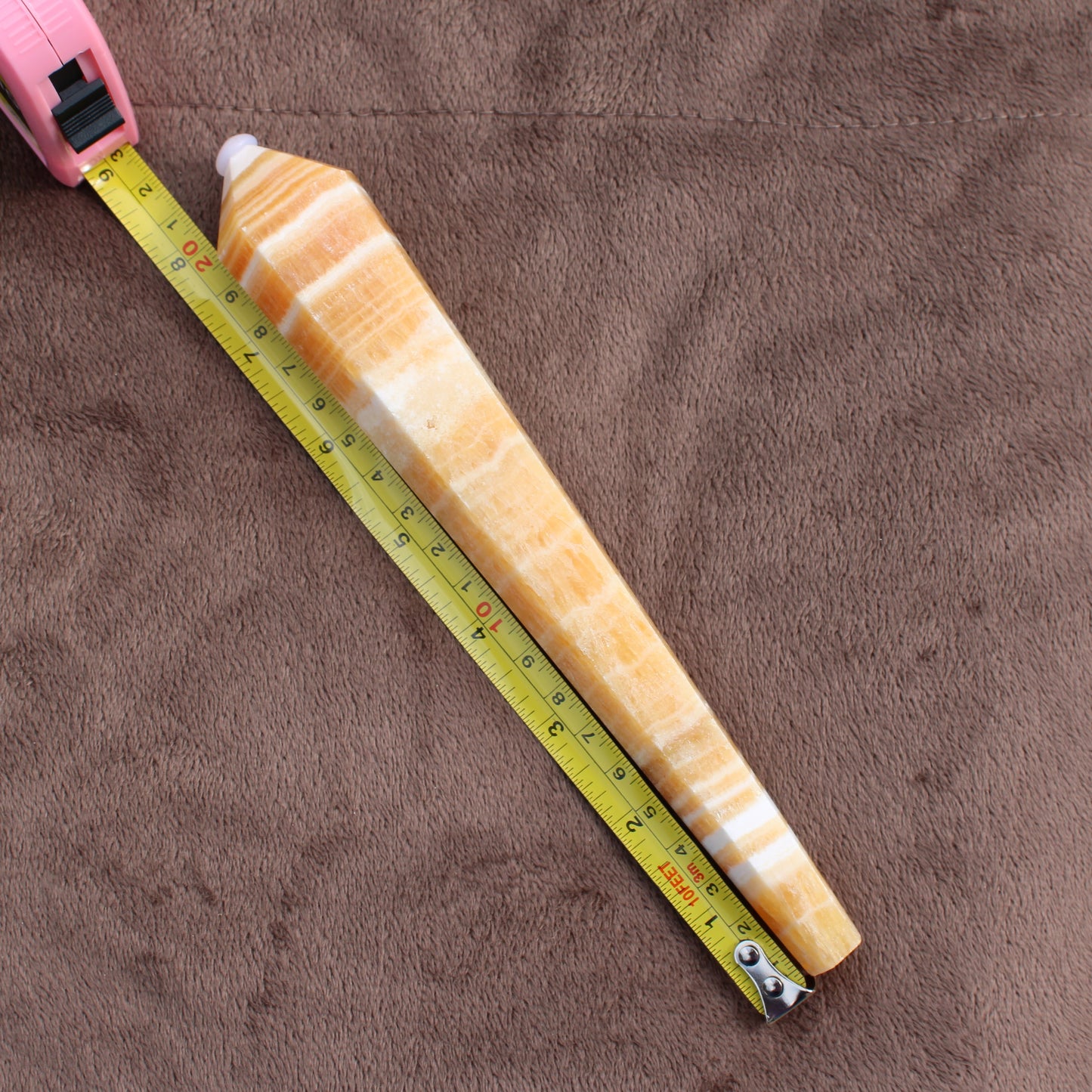 Honey Calcite Wand crystal measuring about 7 inches on a pink tape measure on a brown fabric background