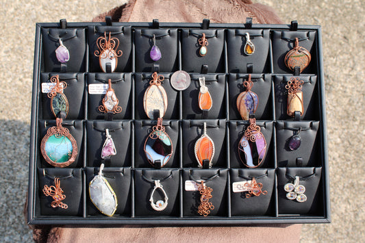 Collection of affordable hand crafted pendants with various stones and intricate wire designs displayed in a black jewelry tray