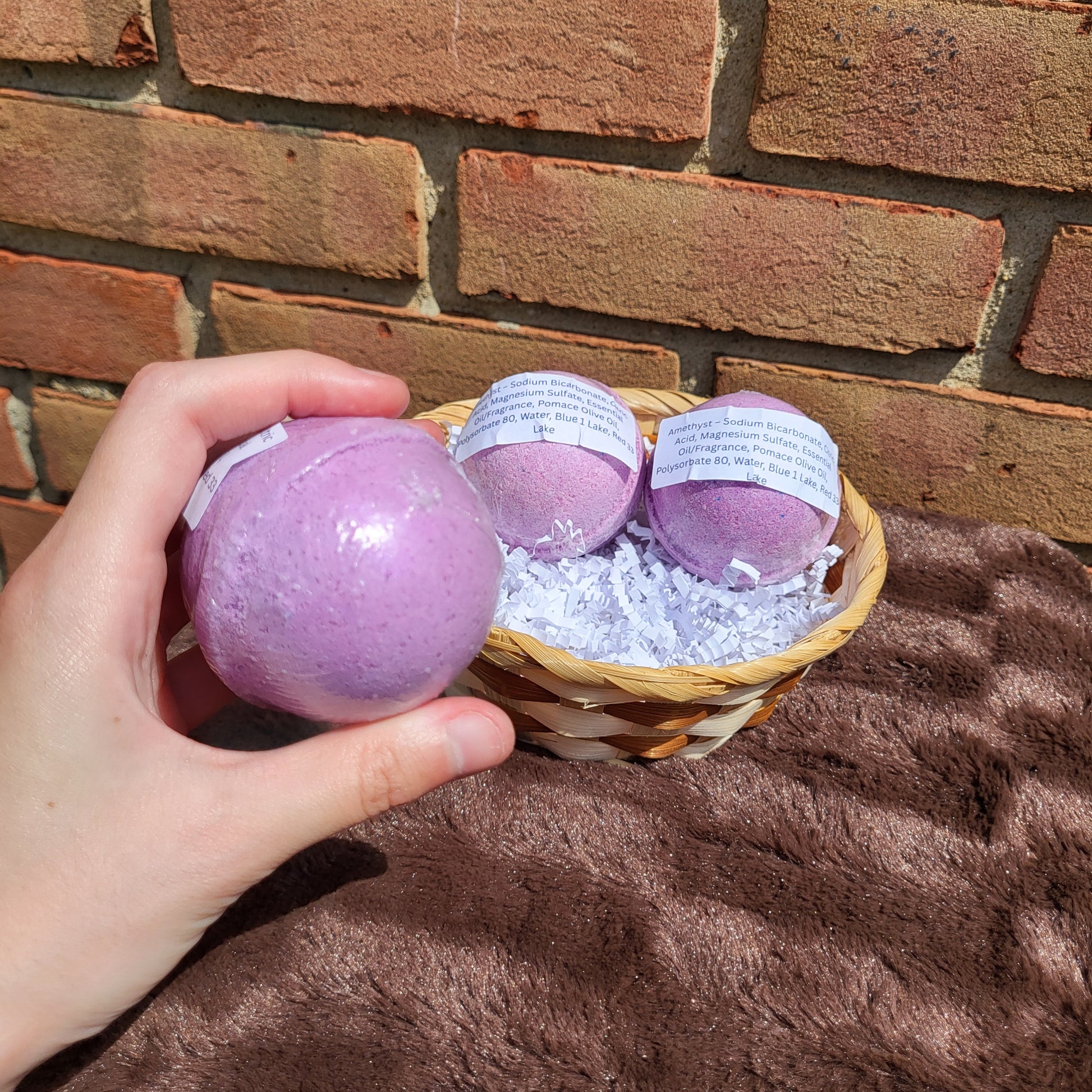Amethyst Bath Bomb in hand with a basket of additional bath bombs on a soft surface