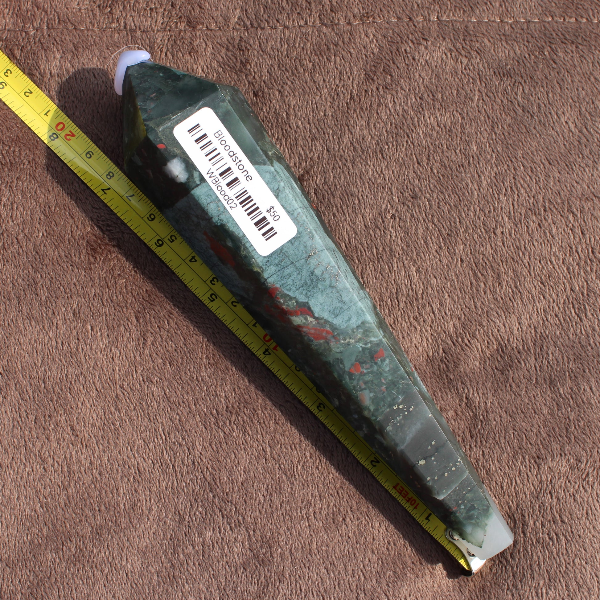 Large Bloodstone Wand crystal with green and red hues displayed with measuring tape on brown fabric