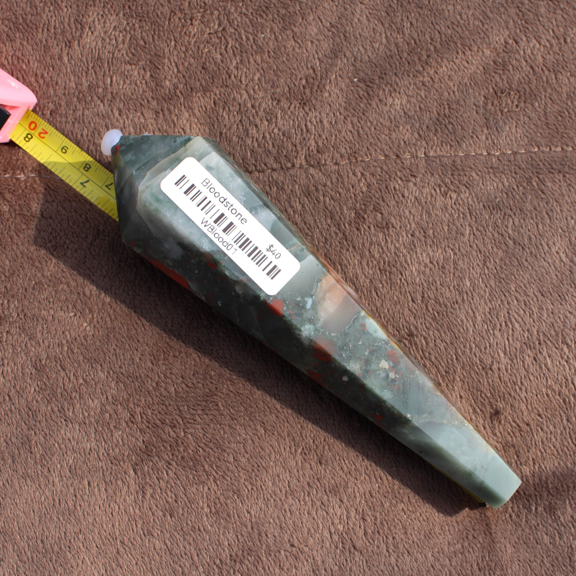 Large Bloodstone Wand crystal with barcode sticker placed next to measuring tape on brown fabric