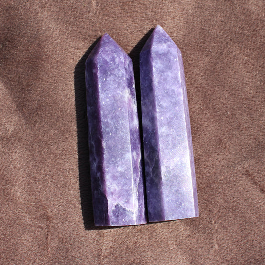 Two polished purple Lepidolite Tower crystals standing upright on a brown fabric background