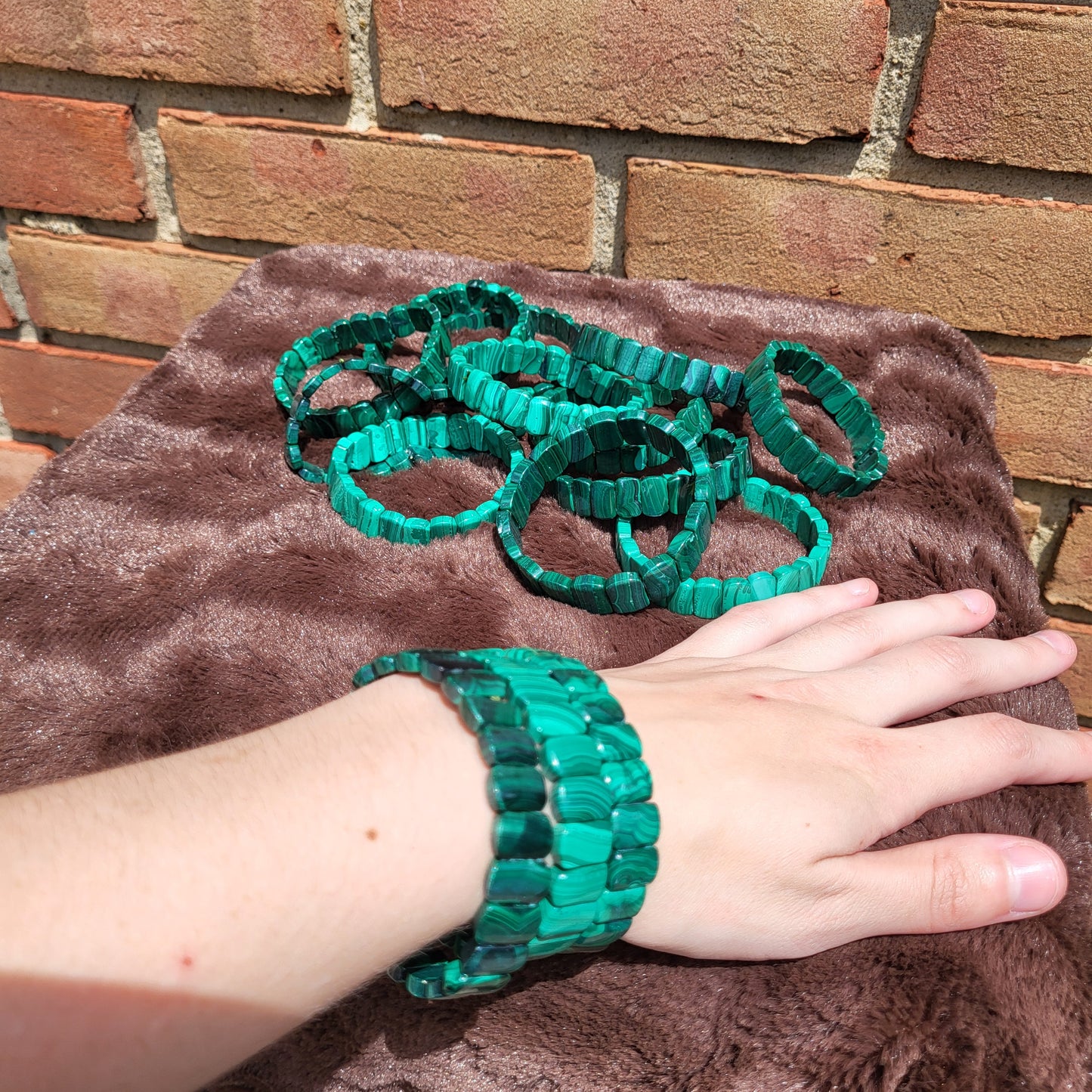 Hand wearing a Malachite Bead bracelet with multiple green bracelets displayed on a brown plush surface