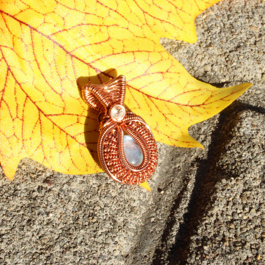 Mini rainbow moonstone copper pendant with intricate design on a vibrant leaf background