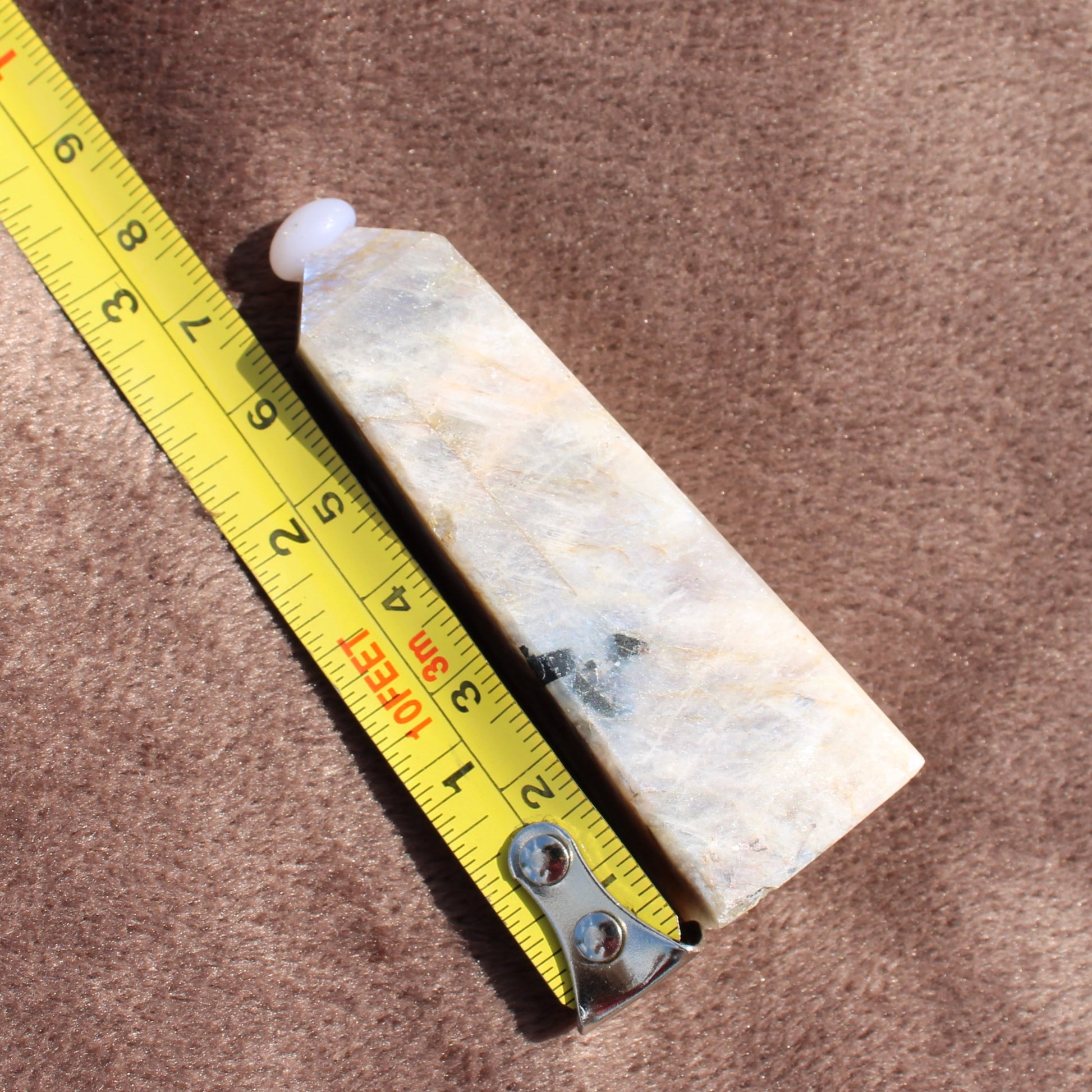 Moonstone Tower crystal from Different Branches LLC next to a yellow measuring tape on a brown surface