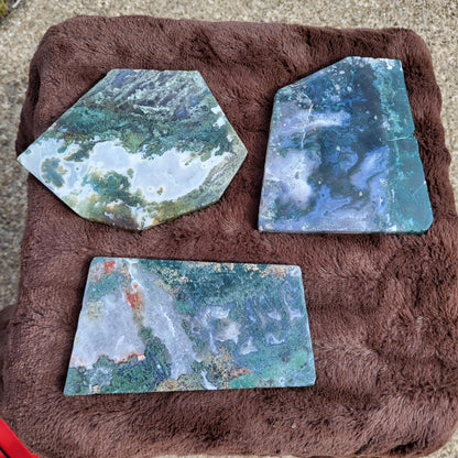 Three Moss Agate Slab pieces with unique green and blue patterns displayed on a textured surface