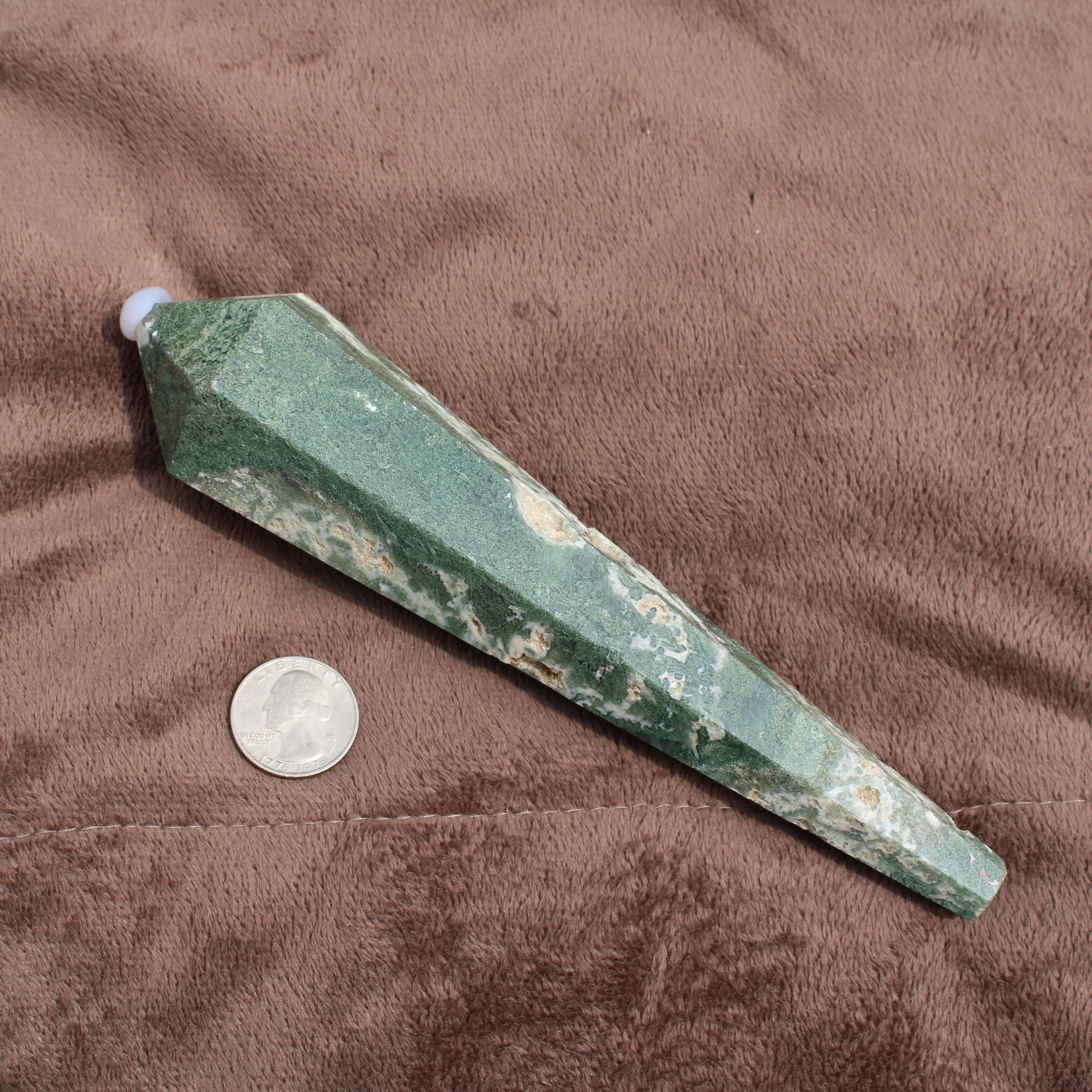 Green and white polished Moss Agate Wand crystal next to a quarter for size comparison