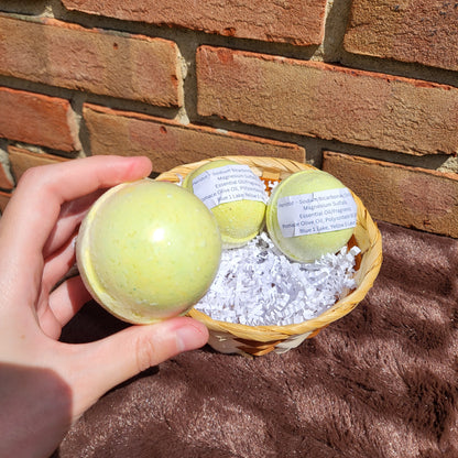 Hand holding a vibrant Peridot Bath Bomb with two more in a basket filled with decorative shreds