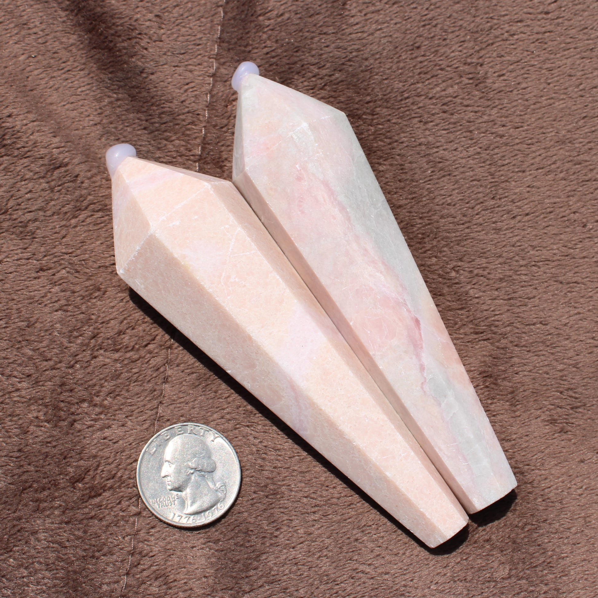 Two polished Pink Opal Wand crystals next to a quarter for size comparison on brown fabric
