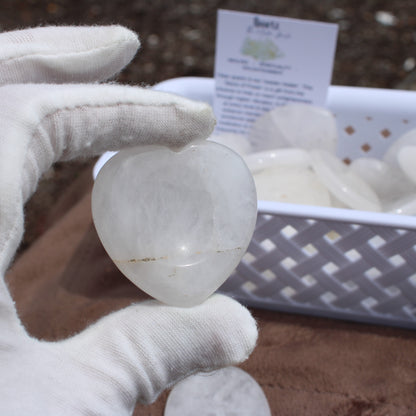 Hand holding a heart-shaped Quartz Worry Stone with white gloves in front of a basket of similar stones