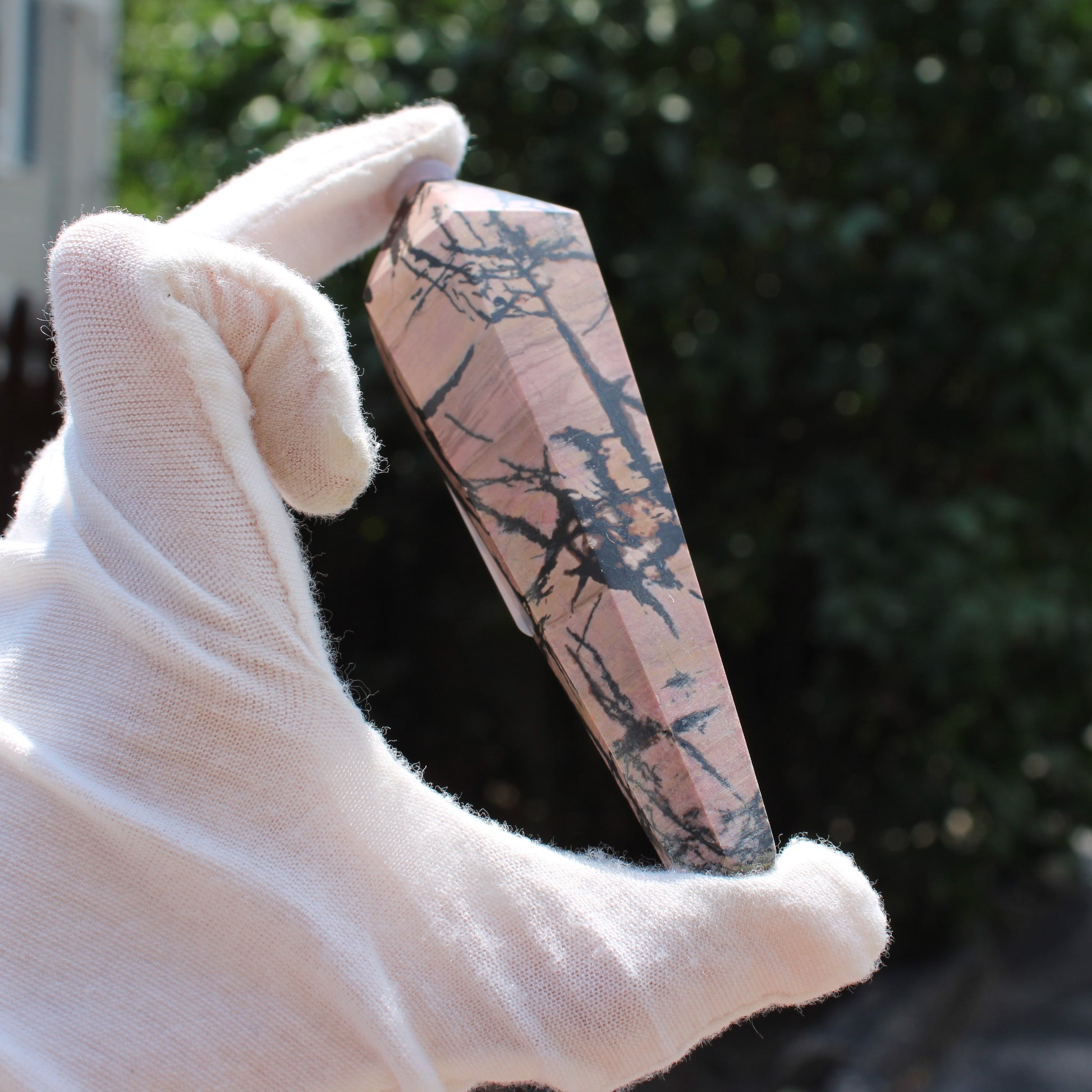 Hand holding a polished Rhodonite Wand crystal with black veining against a blurred natural background
