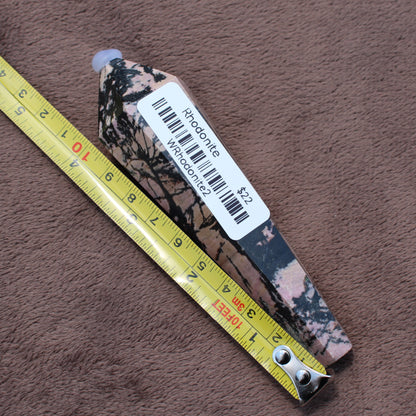 Rhodonite Wand crystal from Different Branches LLC placed next to a yellow measuring tape on a brown surface