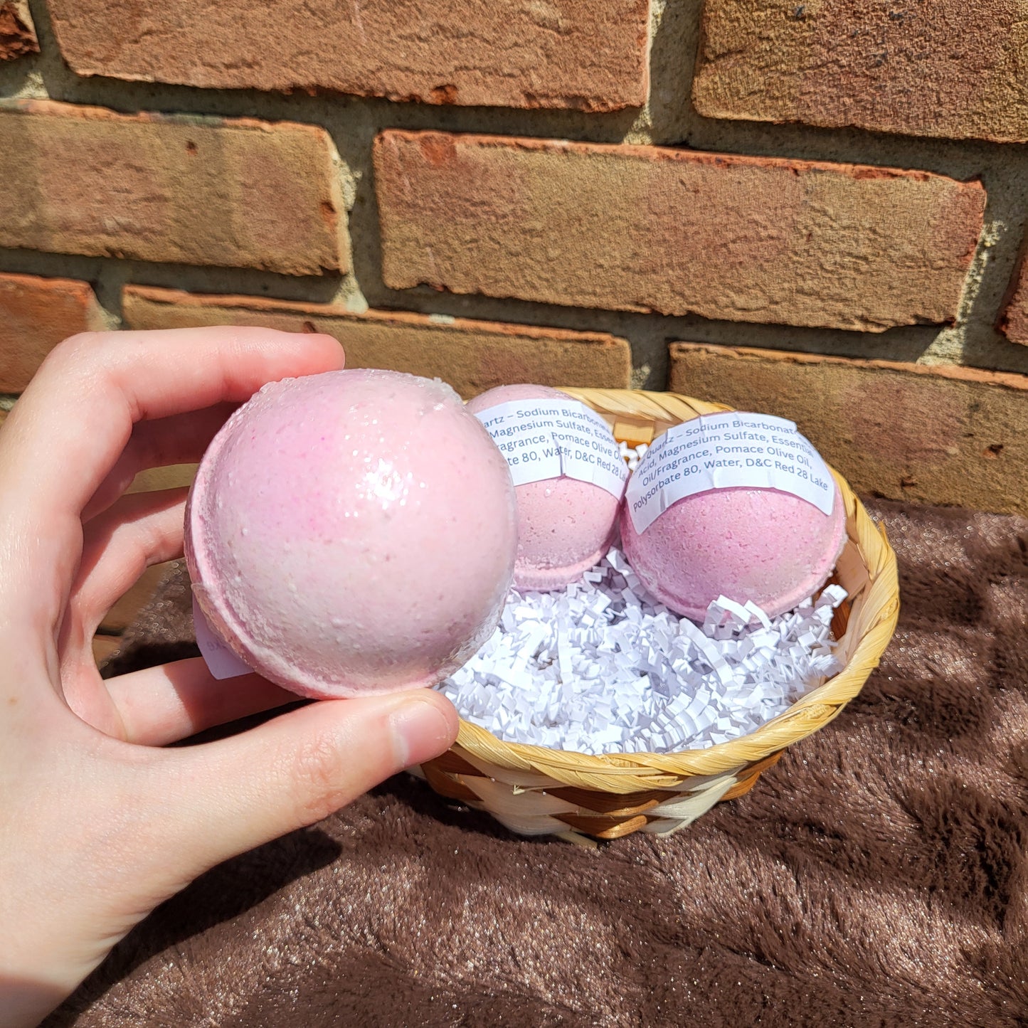 Hand holding a Rose Quartz Bath Bomb with two more in a wicker basket filled with shredded paper