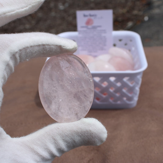 Hand wearing white glove holding a polished Rose Quartz Worry Stone with a basket of similar stones in the background