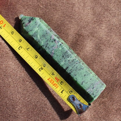 Green and purple Ruby Zoisite Tower crystal point next to a yellow measuring tape on a brown surface