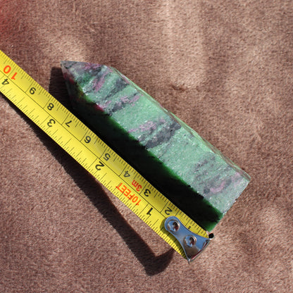 Ruby Zoisite Tower crystal with green and purple hues next to a yellow measuring tape on brown fabric