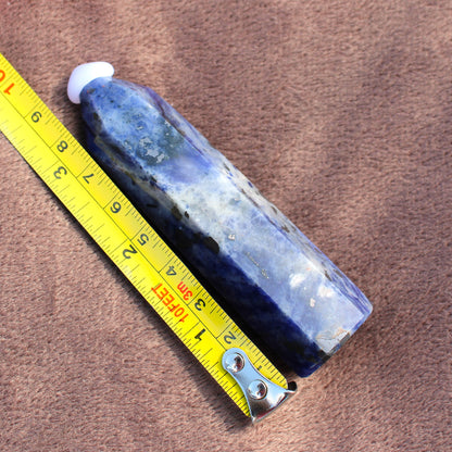 Blue and white polished Sodalite Tower crystal next to a yellow measuring tape on brown fabric