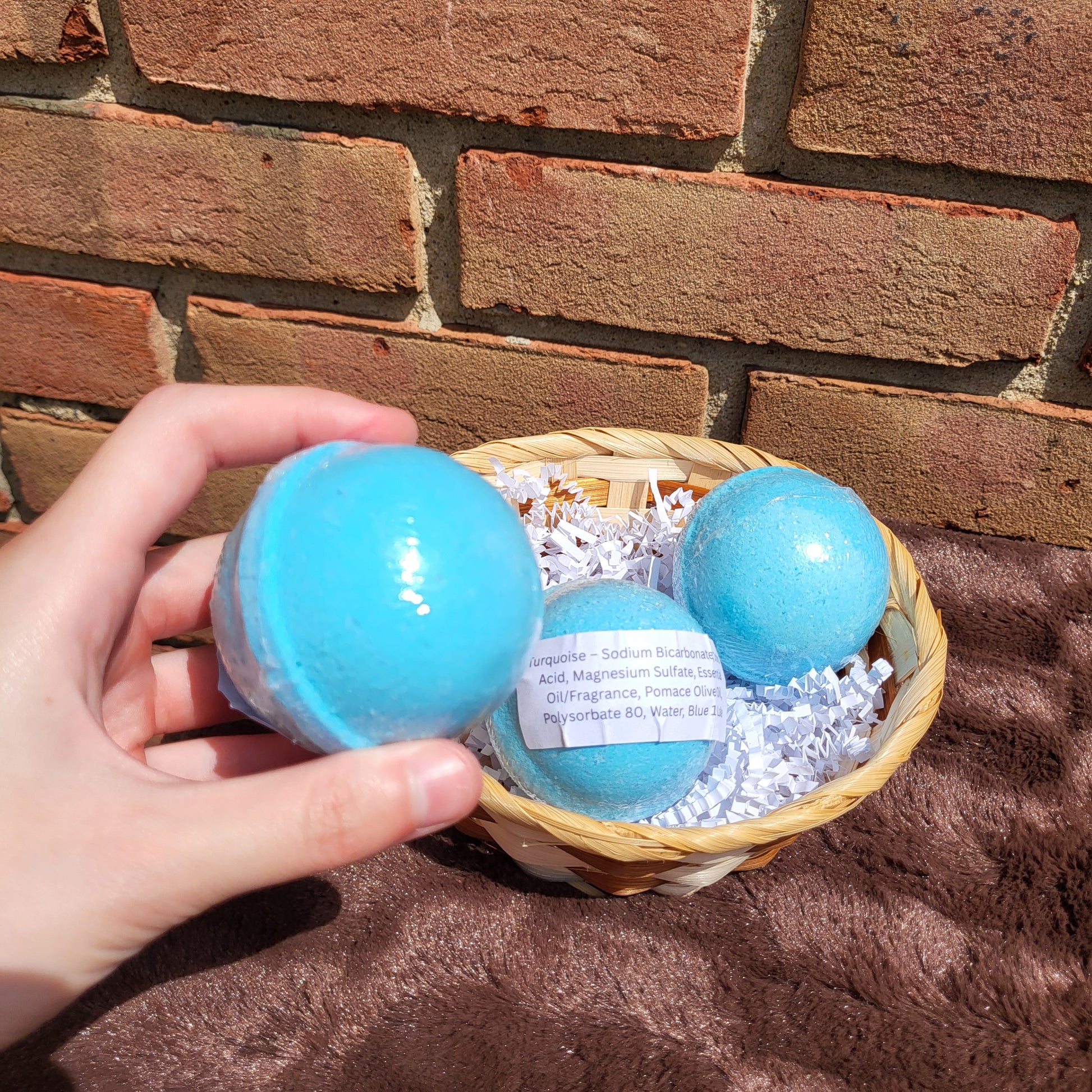 Hand holding a vibrant Turquoise Bath Bomb next to a basket of more bath bombs.